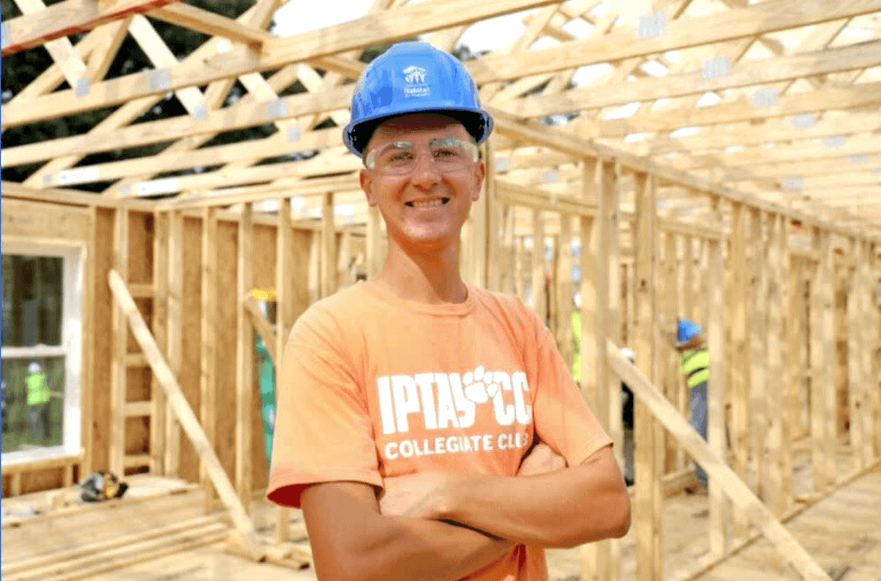 Charlie Rohaley, the Homecoming Coordinator of the Clemson Habitat Student Chapter, standing confidently at a Habitat for Humanity build site. He is wearing a blue Habitat hard hat, safety glasses, and an orange IPTAY Collegiate Club t-shirt, with the woo