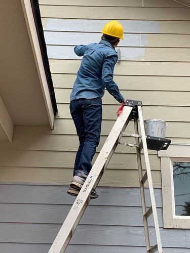 Man in a yellow hard hat on a ladder painting a house