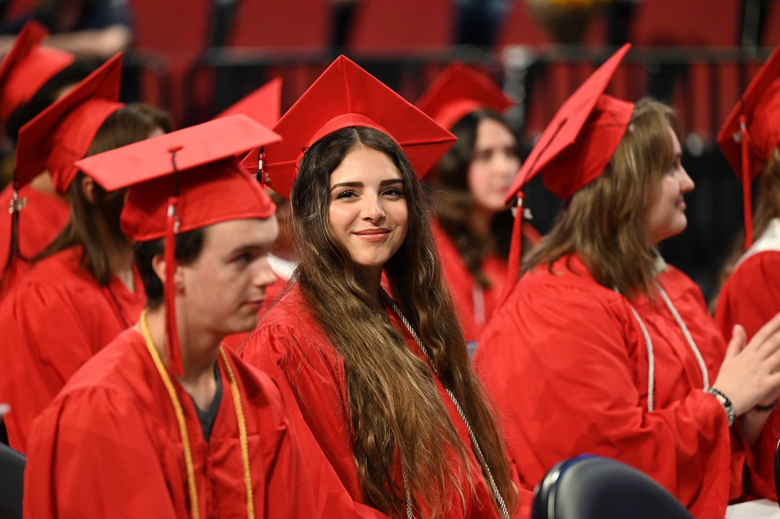 Photo of a young girl dressed in a red cap and gown sitting in the graduation crowd, looking directly at and smiling at the camera.