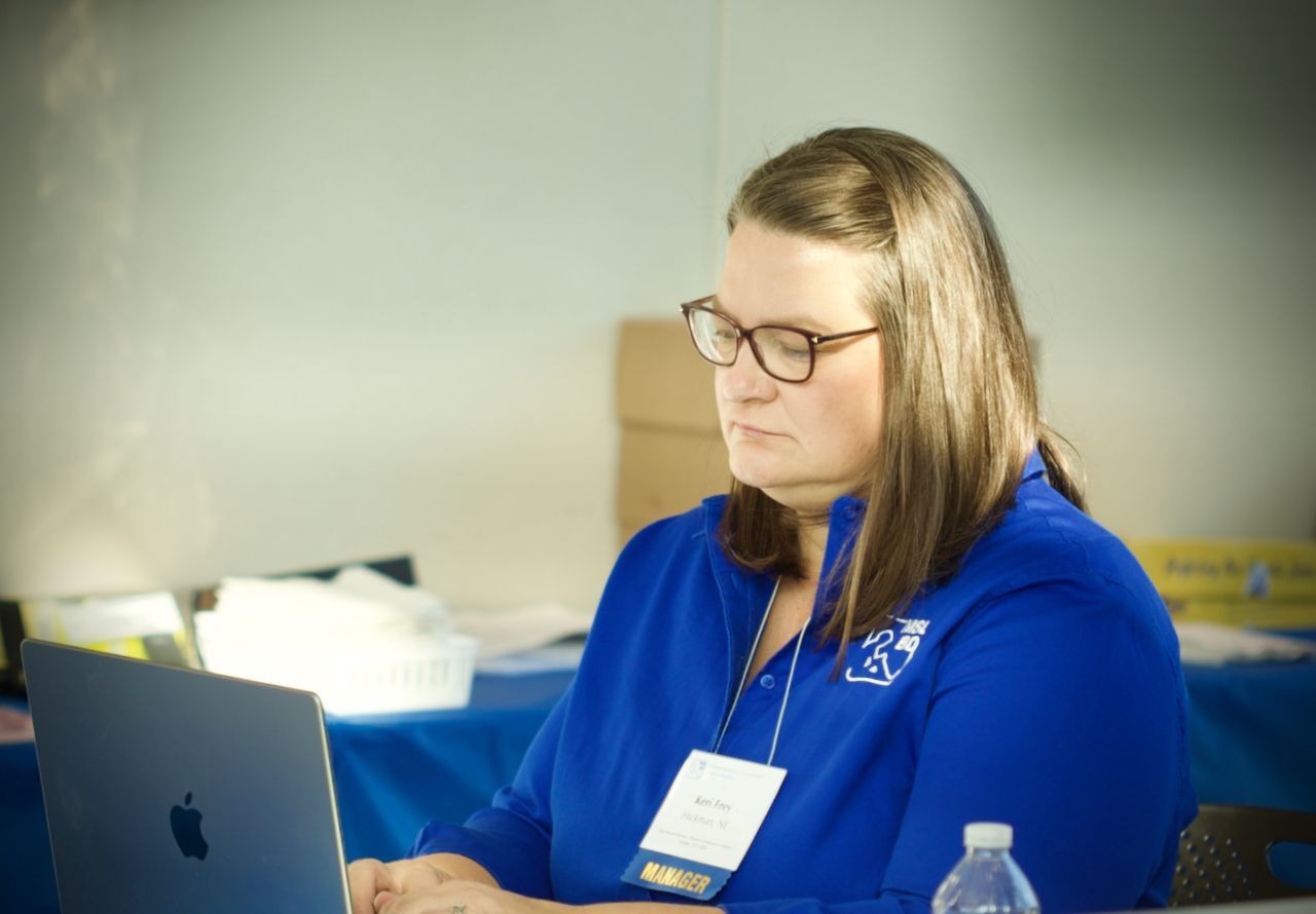 A woman with glasses, Keri Frey, a manager, wearing a blue shirt, works on a laptop at a table with a blue tablecloth.