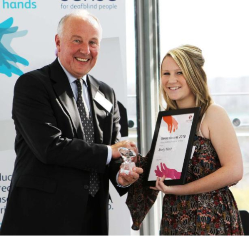 A picture of Molly Watt accepting her Young Deafblind Person of the Year Award. Watt is shaking the hands of a man while she holds her plaque and he holds her award.