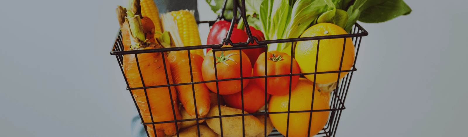 Foto de una canasta hecha de alambre negra y llena de frutas y verduras. El fondo es gris ligero. La canasta contiene zanahorias anaranjadas, maíz amarillo, jitomates rojas, papas morenas, lechuga verde, y naranjas.