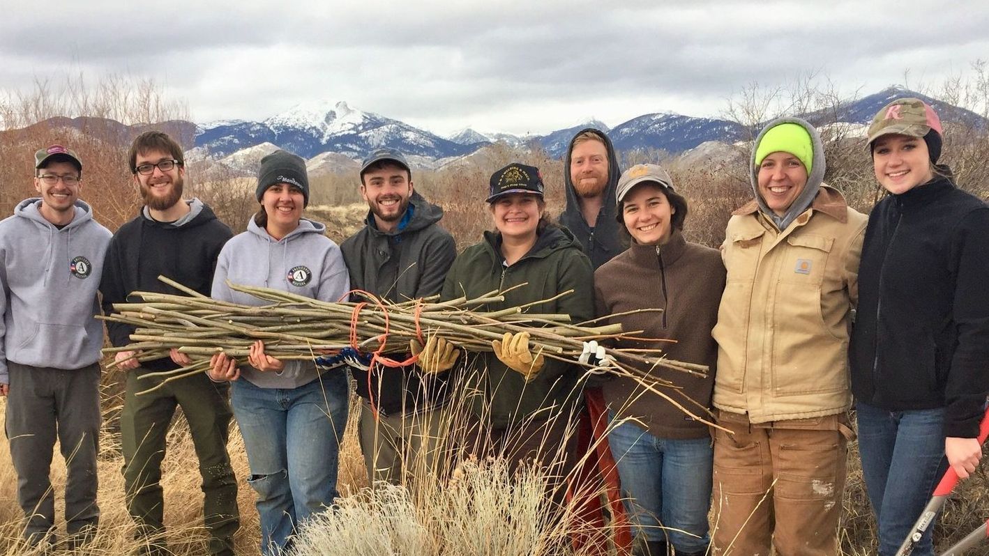 [Image Description: A group of seven Big Sky Watershed Corps members standing together, holding a bundle of sticks. There are snow capped peaks in the background and all the members are cold, but happy!]