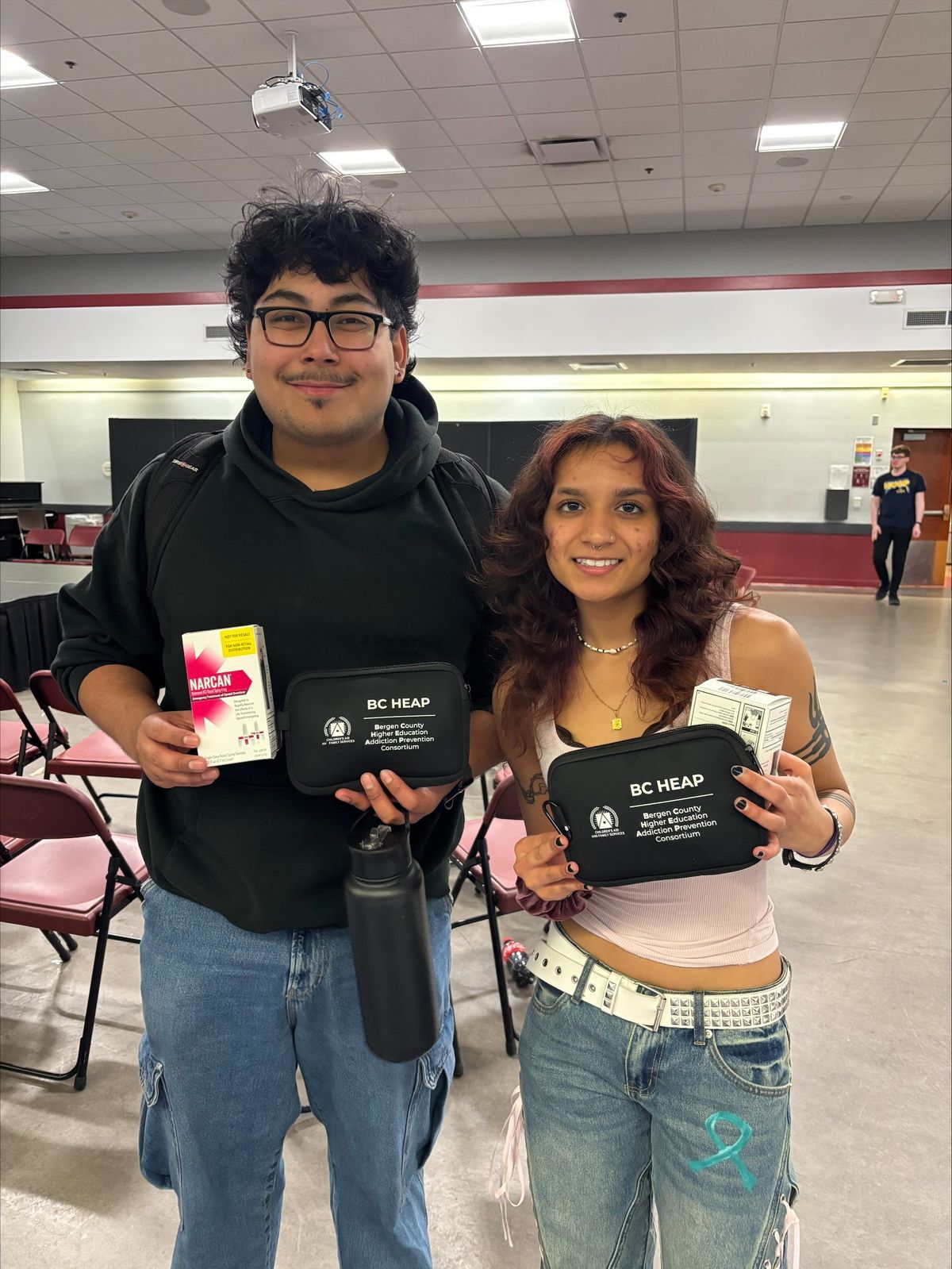 Two individuals stand indoors in a large room with rows of chairs and a ceiling-mounted projector visible above. Each person holds a black case labeled “BC HEAP Bergen County Higher Education Addiction Prevention Consortium,” along with boxes of Narcan.
