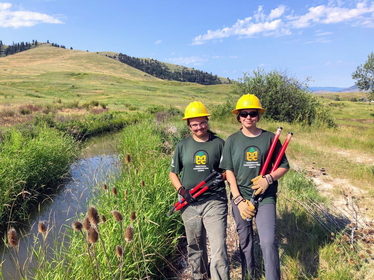 [Image Description: Two crew members holding loppers, standing near a creek with cattails and natural grasses.]
