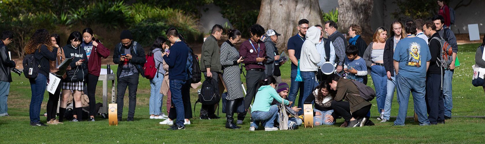 A group of people gathered on a grassy field, some standing and chatting, others looking at electronic devices.