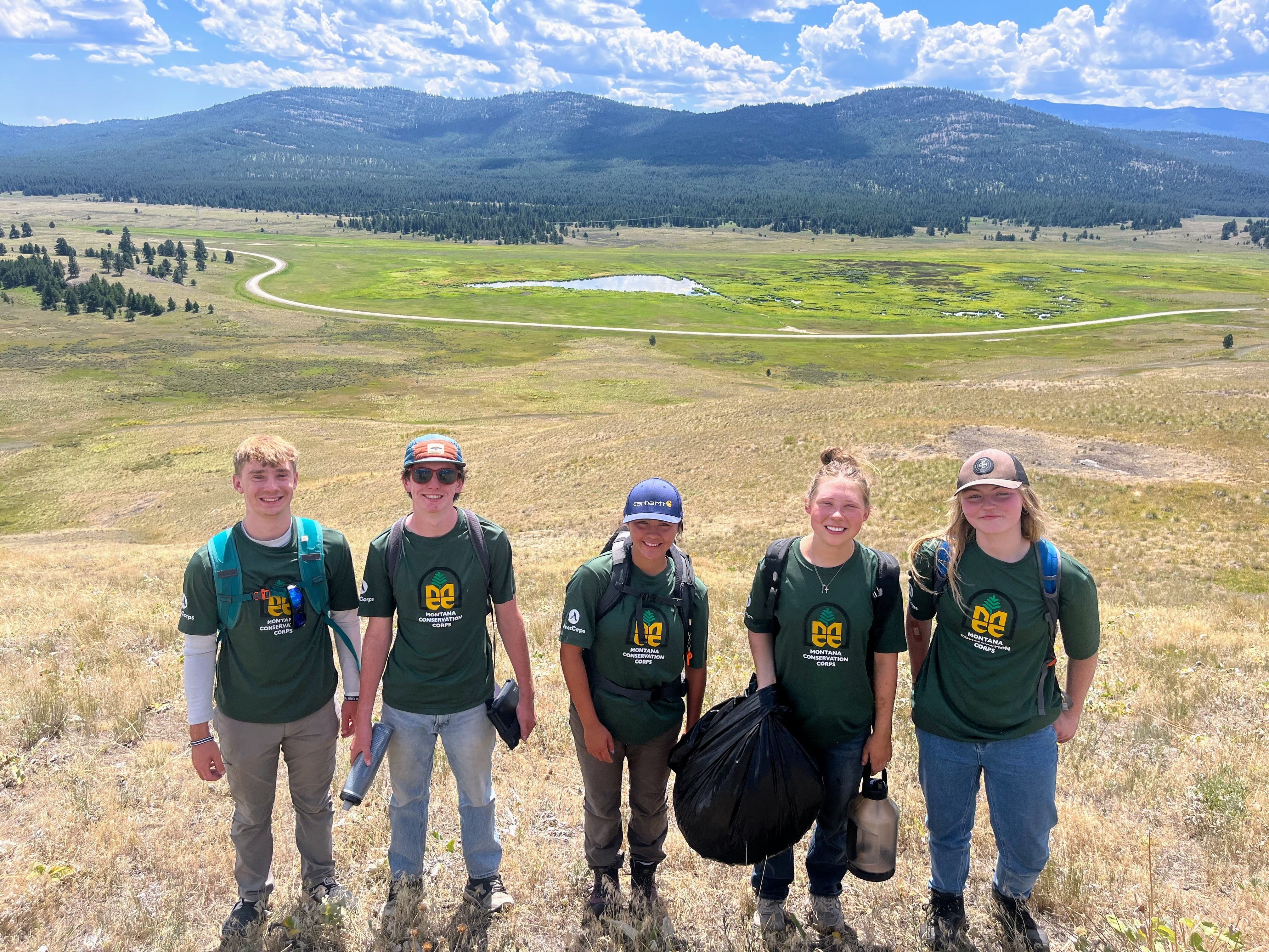 [Image Description: Eight MCC members standing together on a trail wearing their uniforms and helmets, holding shovels and pulaskis. Five of the members are youth, standing alongside their expedition leaders.]