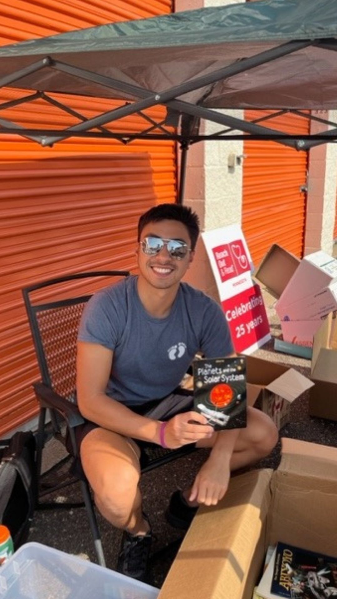 Volunteer sits under tent holding up book, sitting between boxes of books