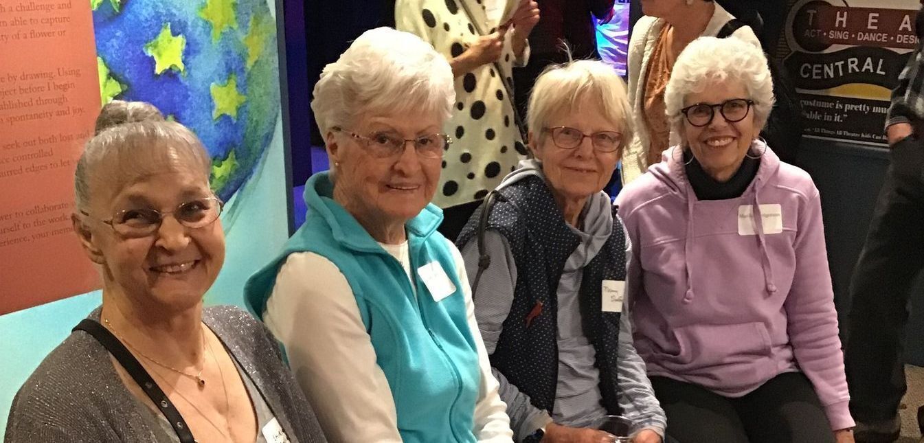 Four older women sitting in an exhibit gallery smiling at the camera.