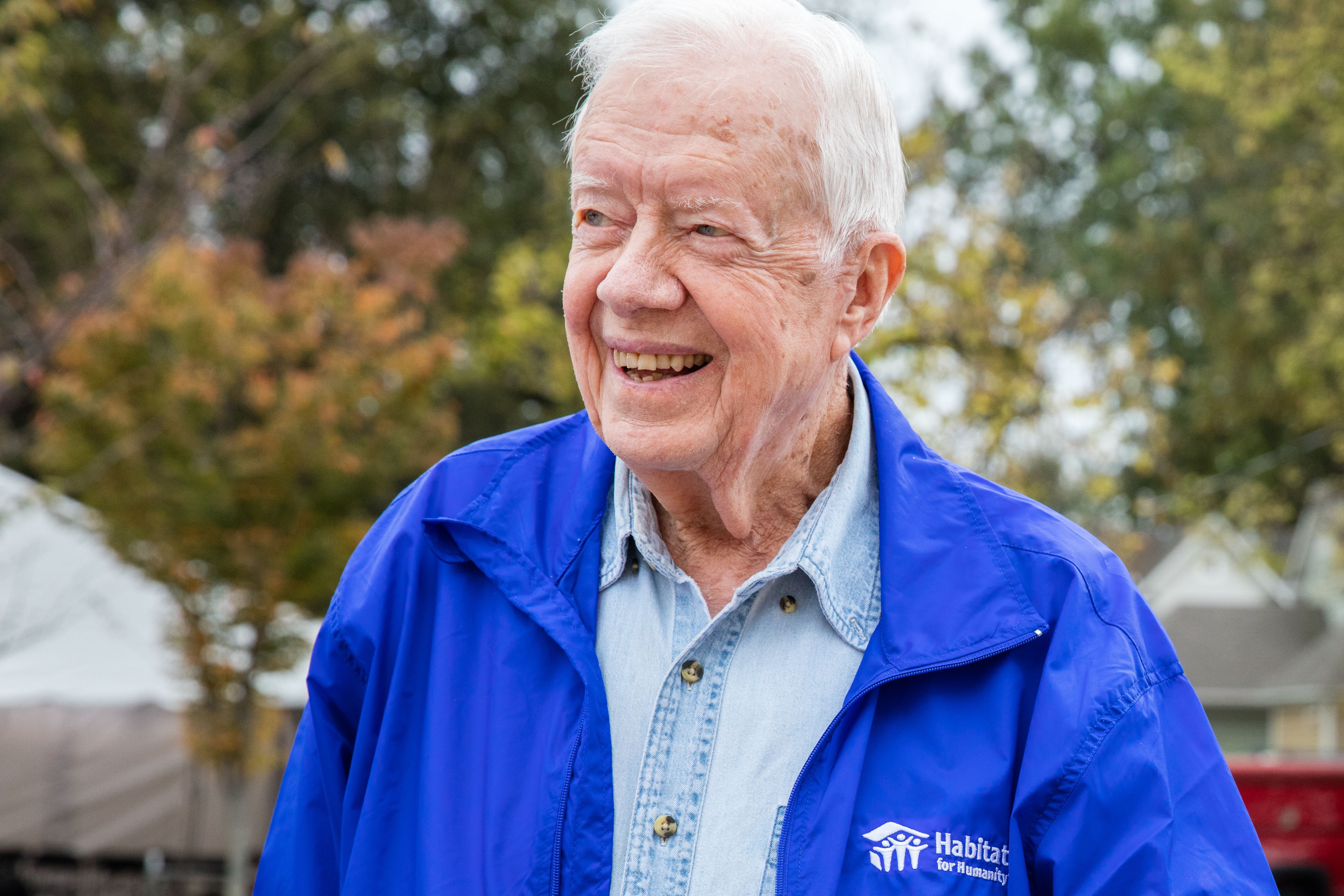 Former President Jimmy Carter smiling outdoors, wearing a blue Habitat for Humanity jacket, symbolizing his decades of volunteerism and leadership in affordable housing efforts.