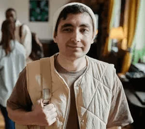 A man with short dark hair is wearing a white knit beanie and a beige quilted vest over a brown shirt. He is holding a strap over his shoulder as if carrying a bag. The background shows an indoor setting with other people and natural light coming through 