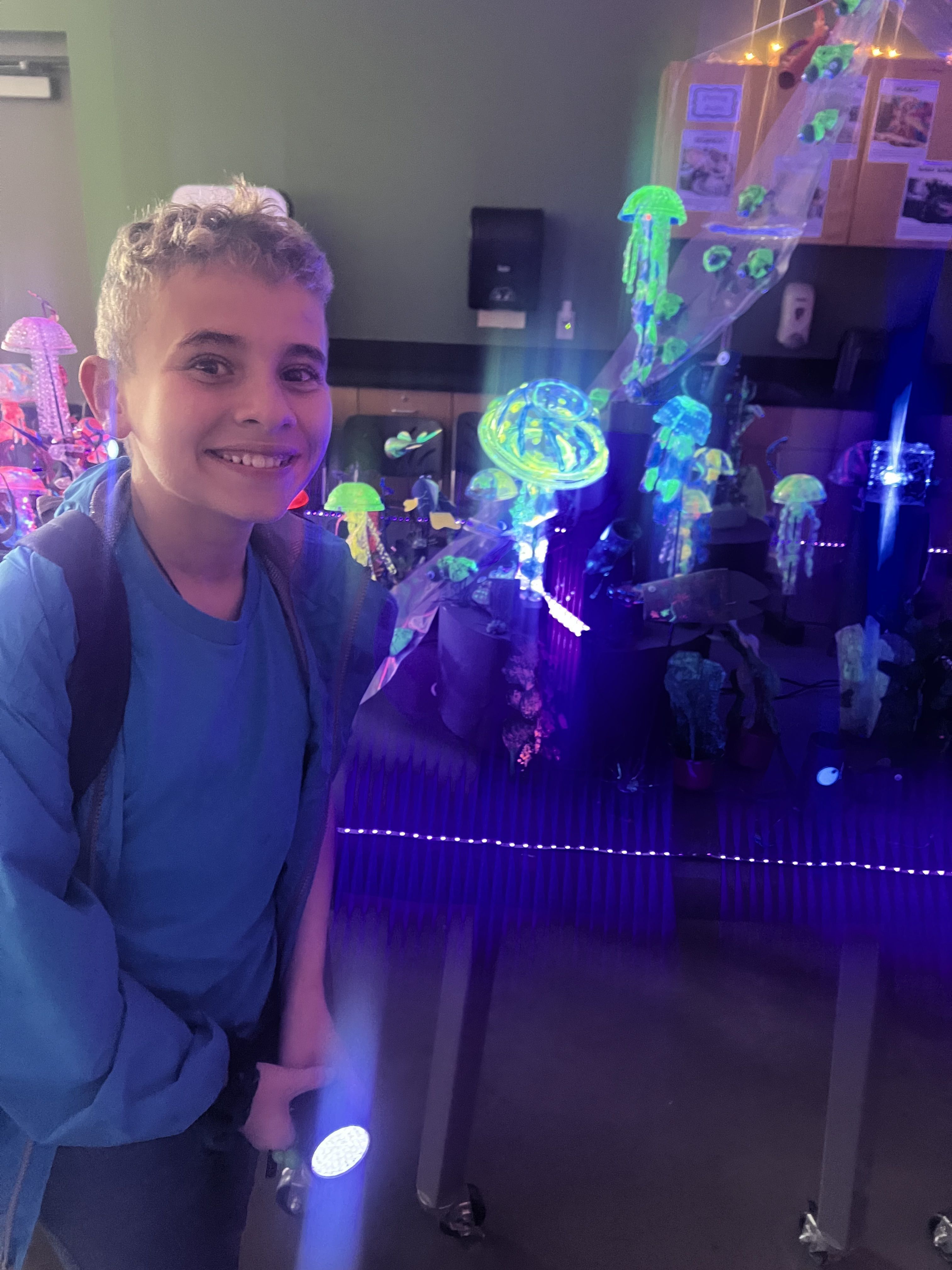 A smiling student with a backpack stands in front of glowing jellyfish displays, capturing the excitement and curiosity sparked by a field trip experience.
