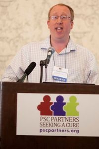 PSC researcher Gideon Hirschfield stands at a podium behind a microphone talking. The podium has a PSC Partners logo on it.