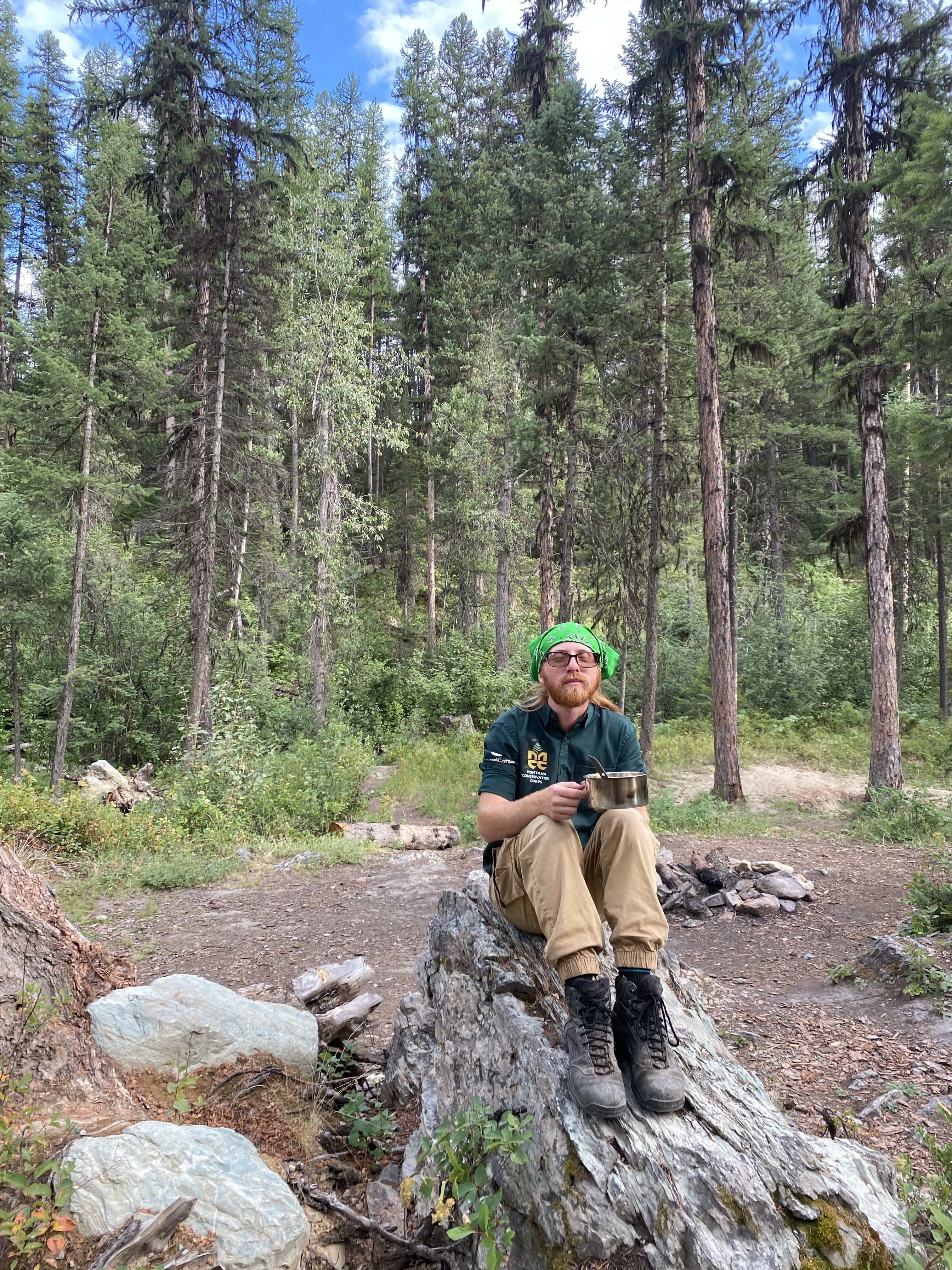 A crew member sits in a campsite, eating from a pot