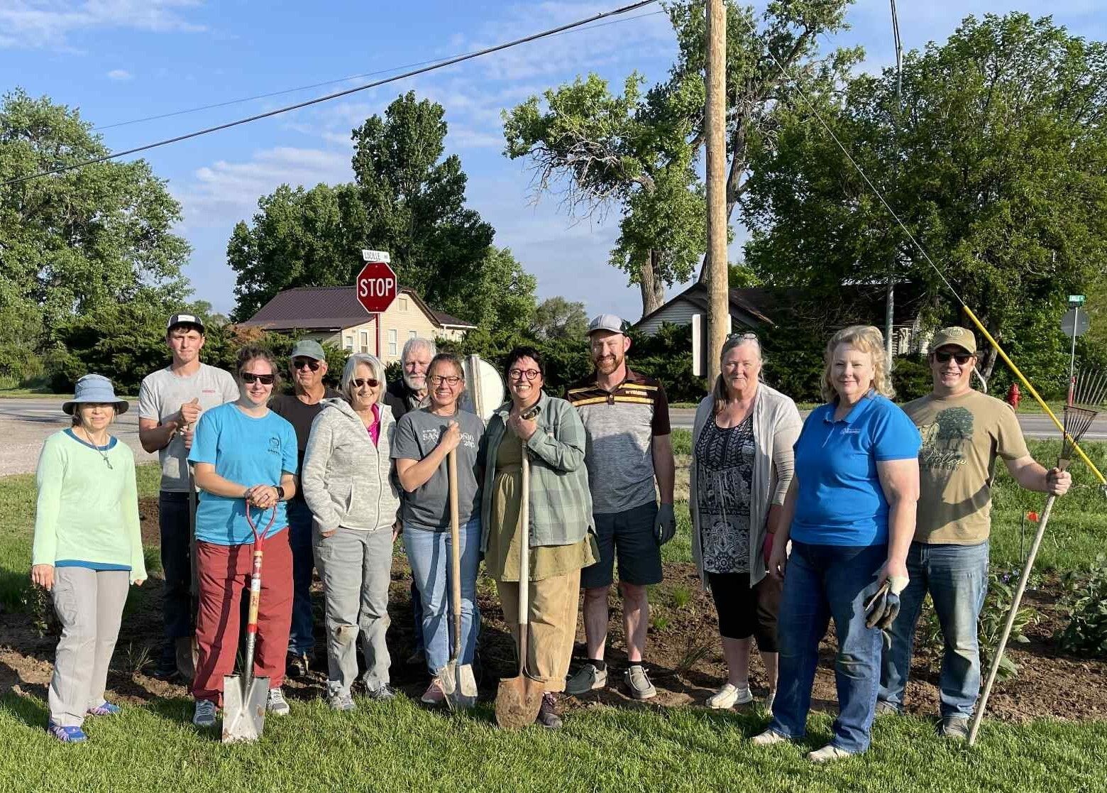 A group of people holding gardening tools pose in front a garden they planted.