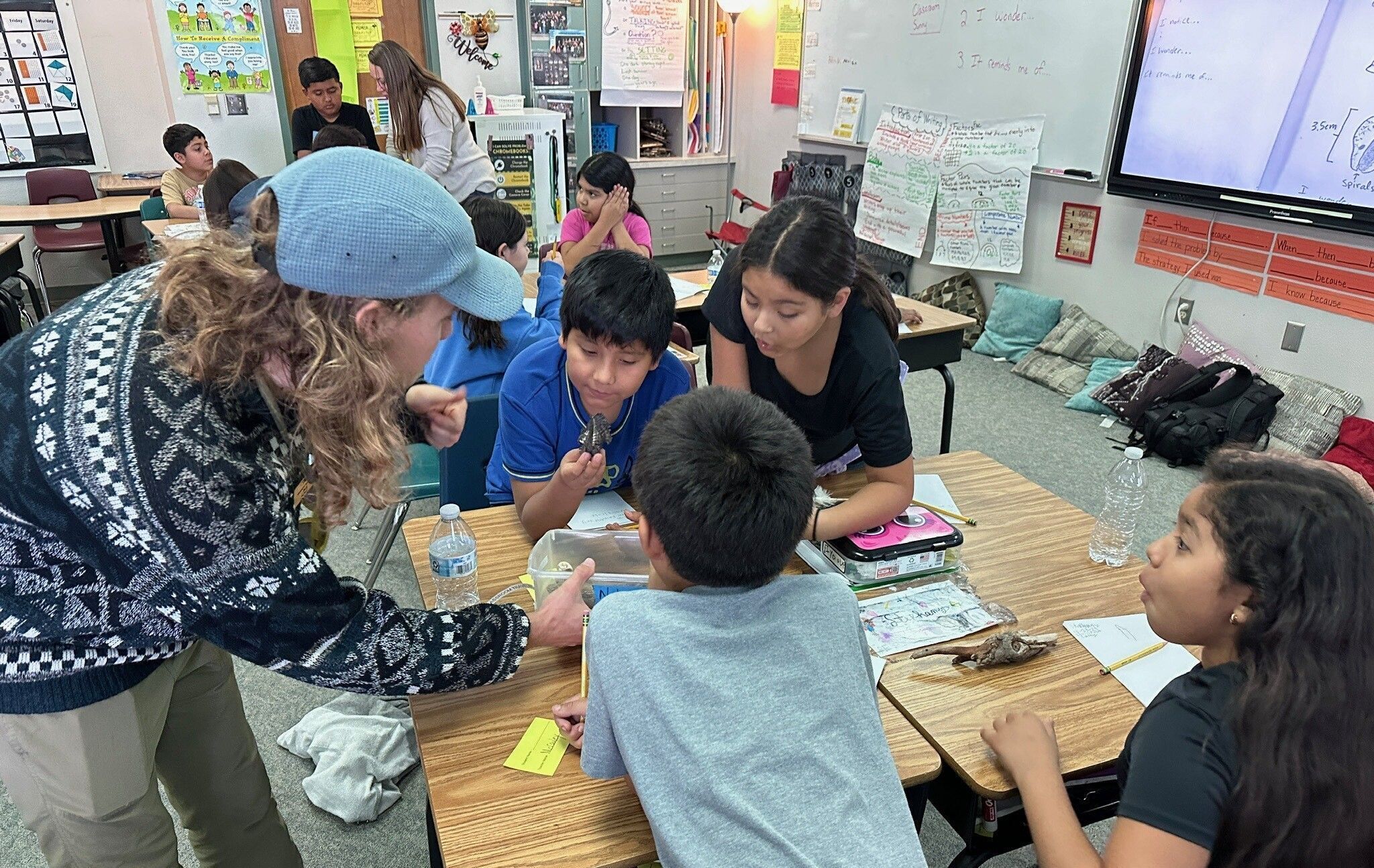 An educator leaning over desks at a school showing 4 students a box of nature discoveries.