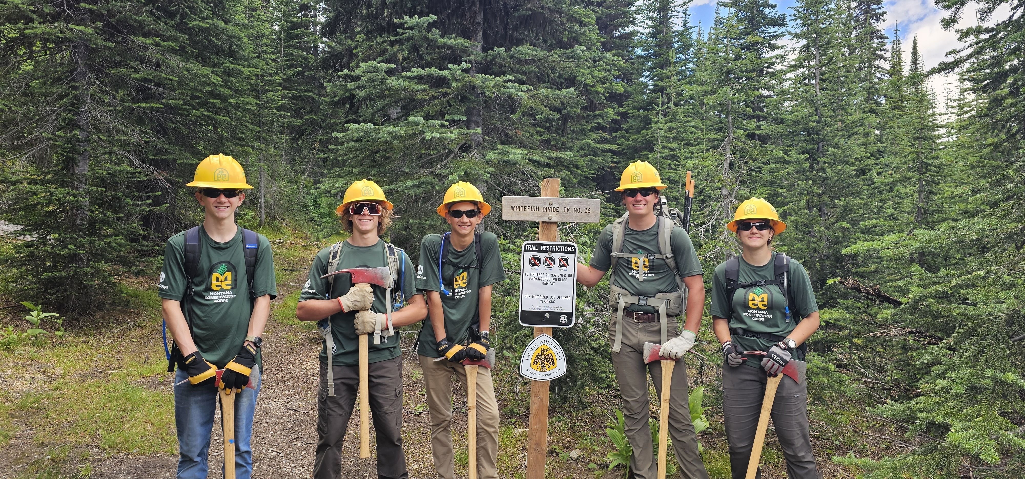 A youth crew stands smiling next to a trail sign