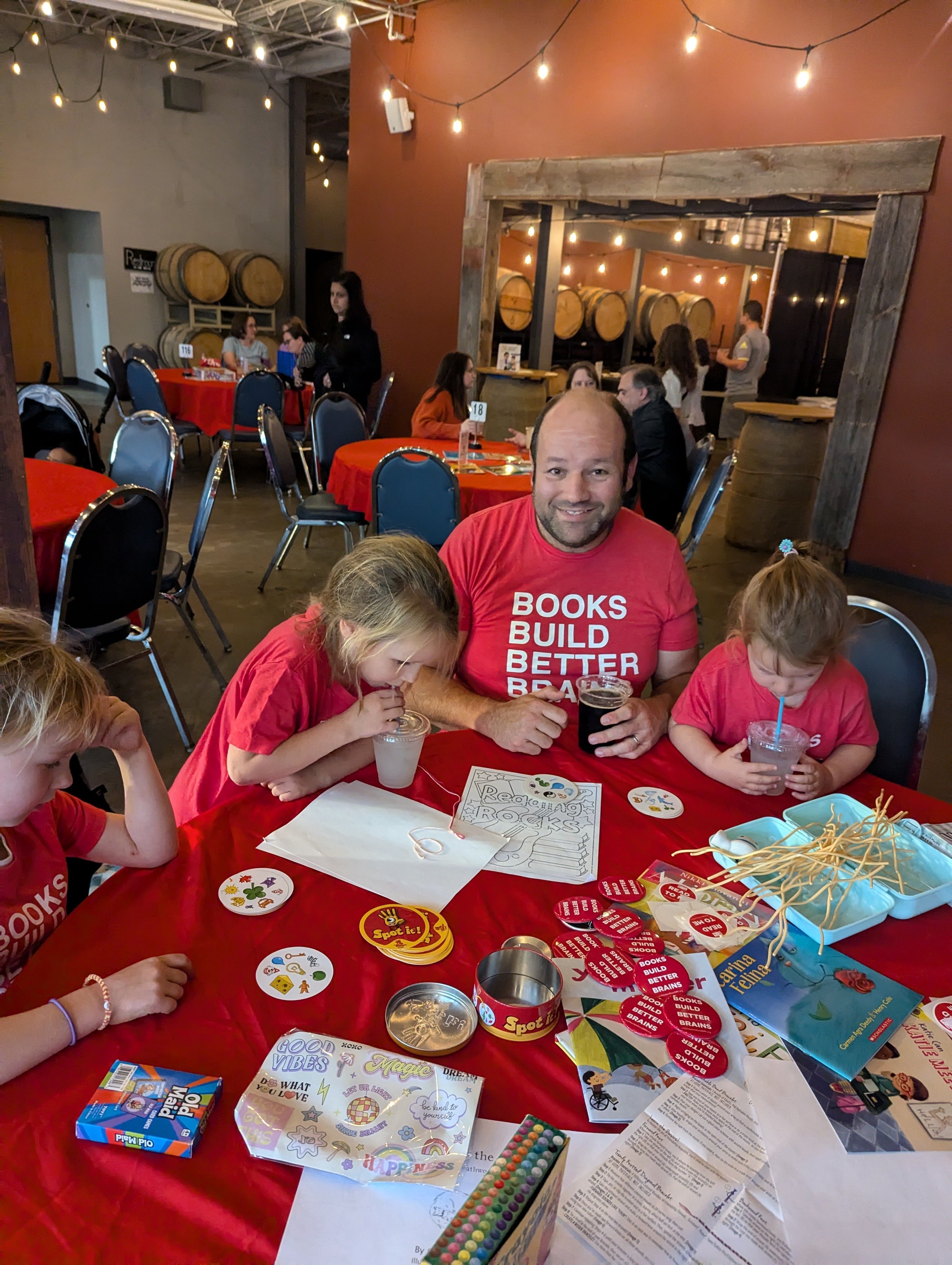 man and three young girls sit at coloring table wearing BOOKS BUILD BETTER BRAINS shirts