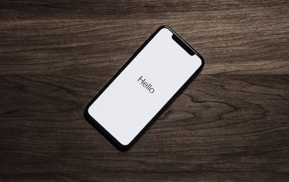 Aerial photo of a smart phone laying on a wooden desk. The phone lays at an angle. The phone shows the word "Hello" in black letters on a white background.
