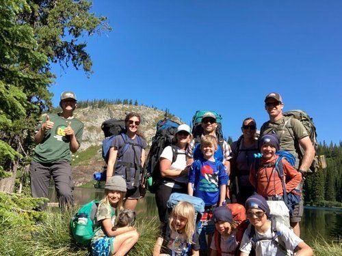 An individual wearing an MCC shirt and hat poses with a group of kids, all wearing backpacks.