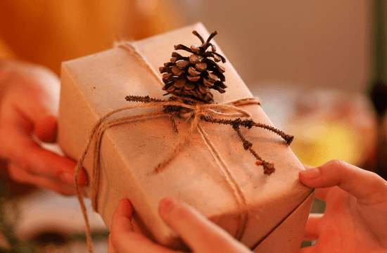 a person holds a gift wrapped in plain brown paper, tied with twine and a pinecone accent