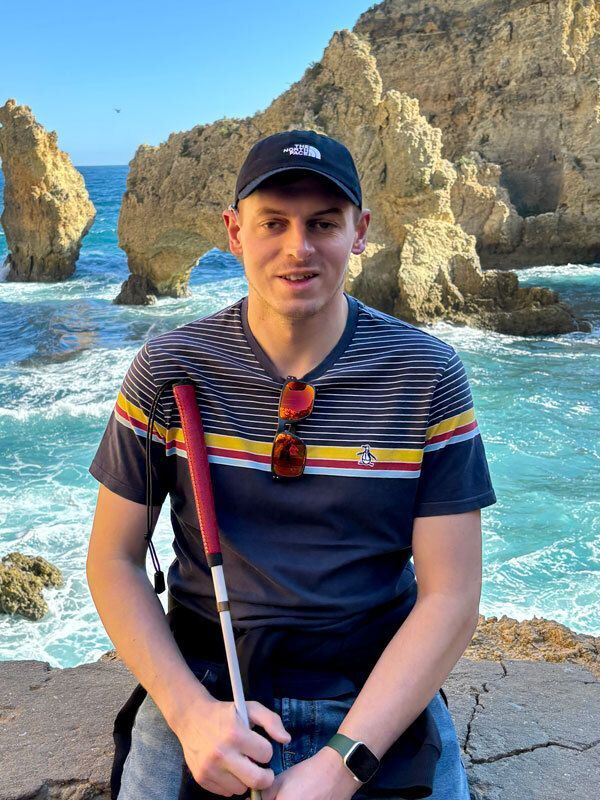 Jeff, smiling, leans on a rock, with the ocean and rock formations behind him. He wears a striped shirt, a baseball cap and holds a mobility cane with a red handle..