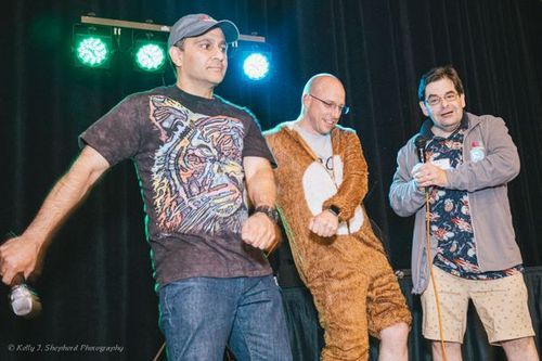 Three men stand in a line and appear to be dancing. One is holding a microphone and wearing a tshirt with a tiger face on it. The man in the middle is wearing a bear costume. 