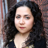 Sofiya Cheyenne, a women with a serious look on her face. She has dark, curly hair and is wearing a patterned blouse. Her head is tilted towards the camera.