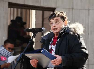 An older woman with short hair and glasses is speaking into a microphone at an outdoor event. She is wearing a black coat with a fur-trimmed hood and holding a blue folder. The setting includes blurred figures in the background near a building with column