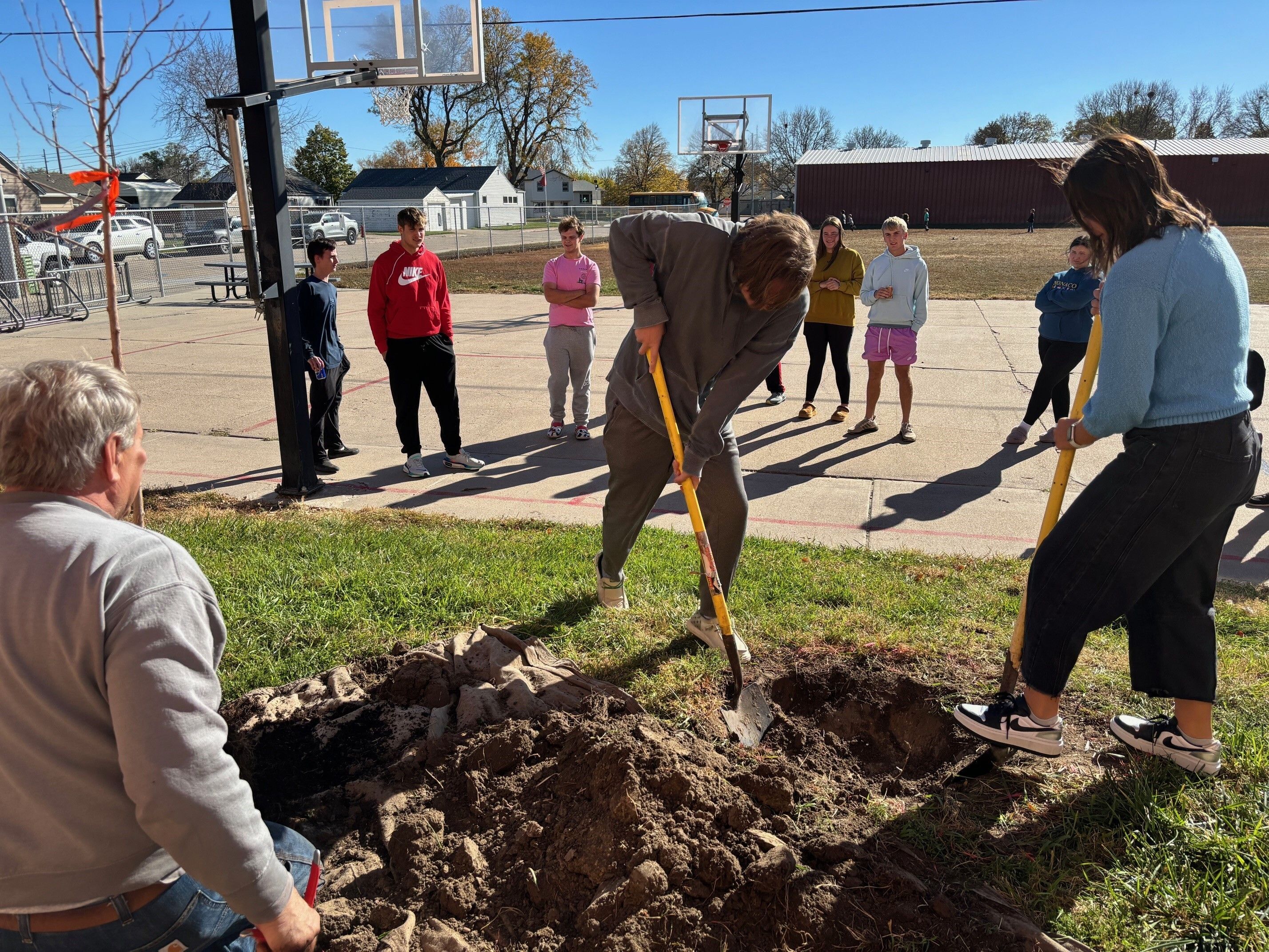 Two women dig a hole for a tree planting while high school students look on