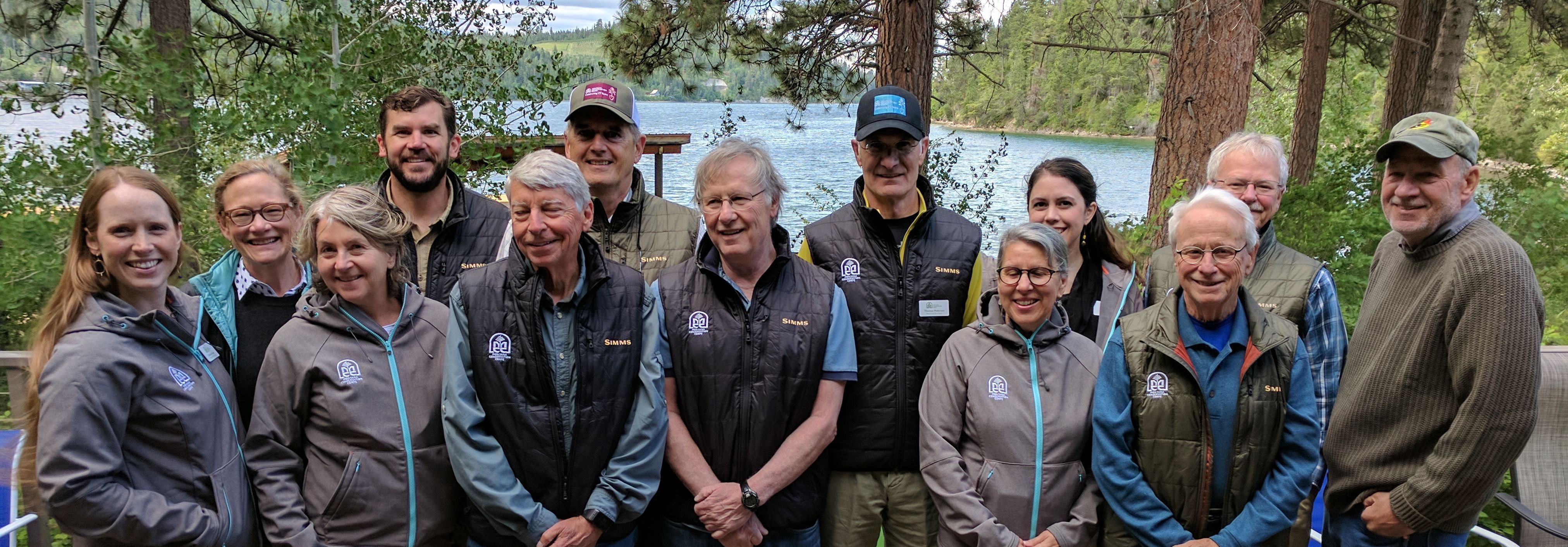 [Image Description: Thirteen members of the Board of Directors standing together on an overlook, wearing their MCC jackets and vests. Behind them is a bright blue lake surrounded by trees.]