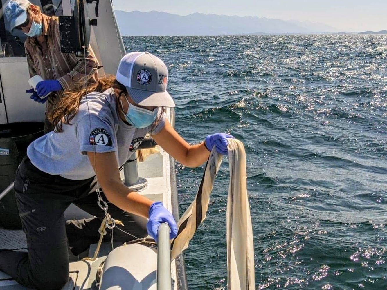 [Image description: a Big Sky Watershed Corps member pulls a net in while on a boat to monitor for Aquatic Invasive Species. They are wearing an AmeriCorps hat, mask, and gloves.]