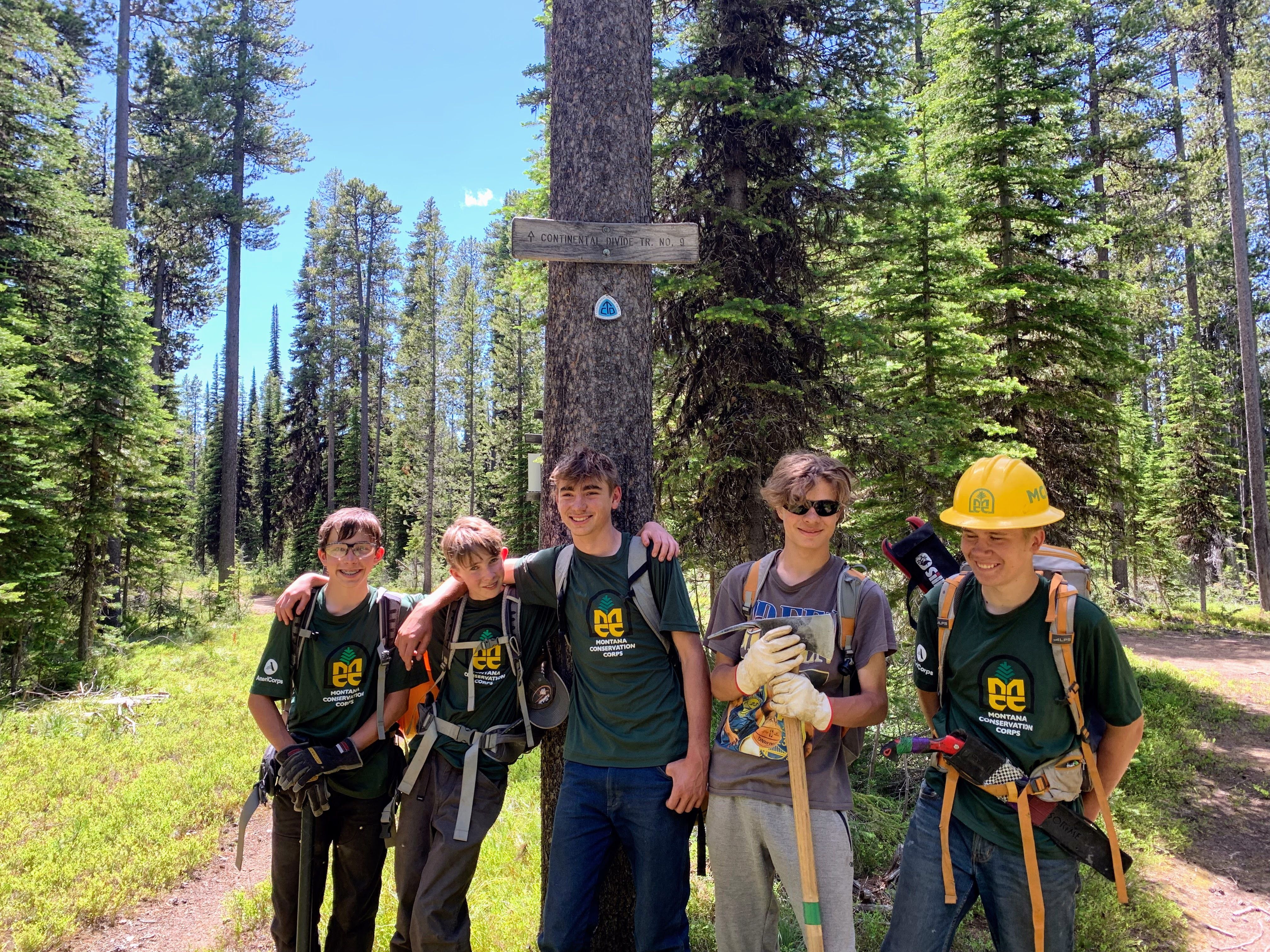 [Image Description: Eight MCC members standing together on a trail wearing their uniforms and helmets, holding shovels and pulaskis. Five of the members are youth, standing alongside their expedition leaders.]