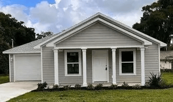 Completed Habitat for Humanity home with gray siding, white trim, and a front porch supported by columns, set against a backdrop of trees and blue sky