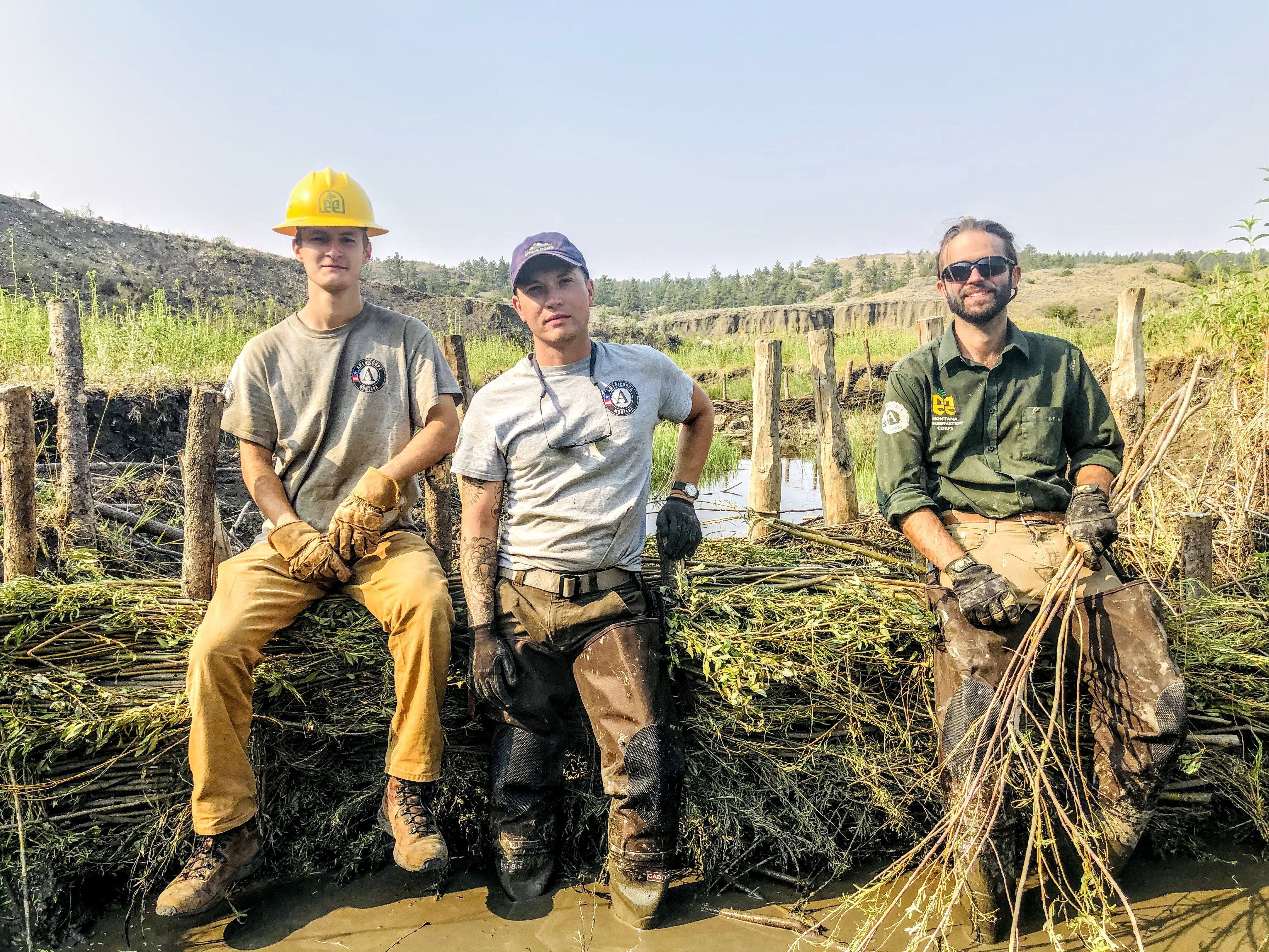 [Image description: Three adult members sit on top of a beaver mimicry dam that they have built to raise the water table and increase watershed resilience.]