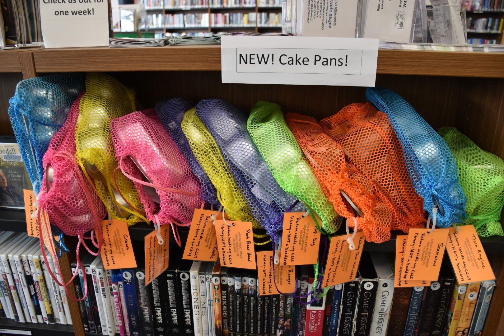 Specialty cake pans in colorful mesh bags on a library book shelf.