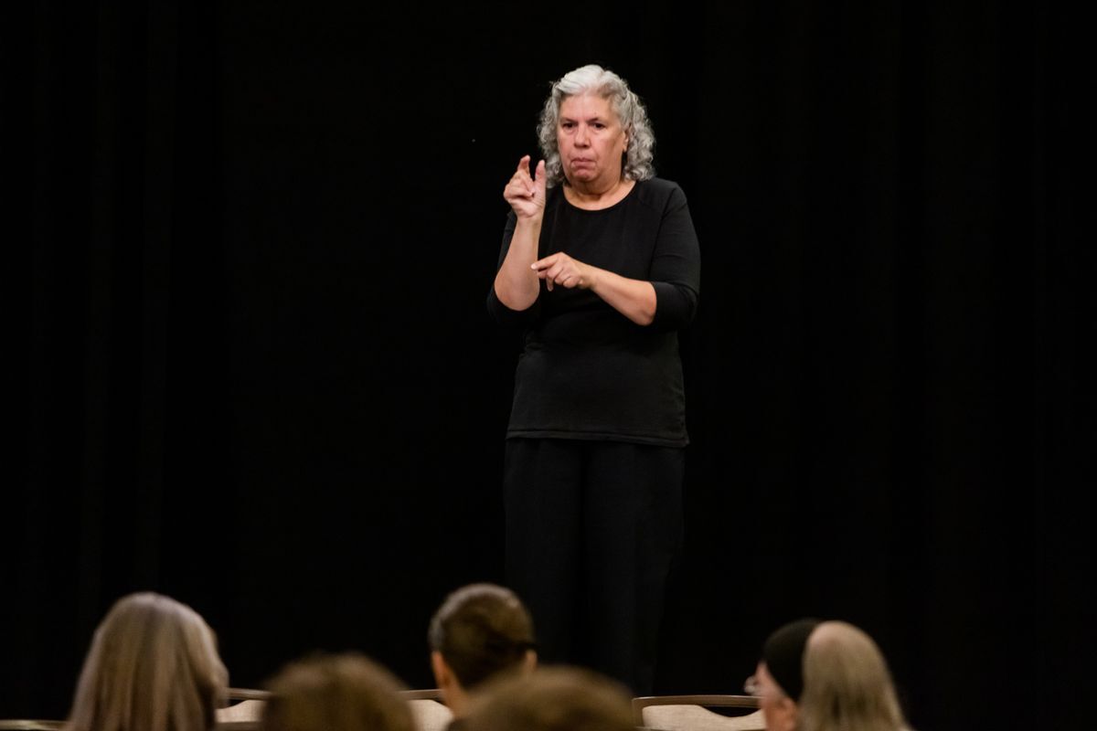 The ASL Interpreter is a white woman who has short grey hair and is wearing all black, standing in front of a dark background. 