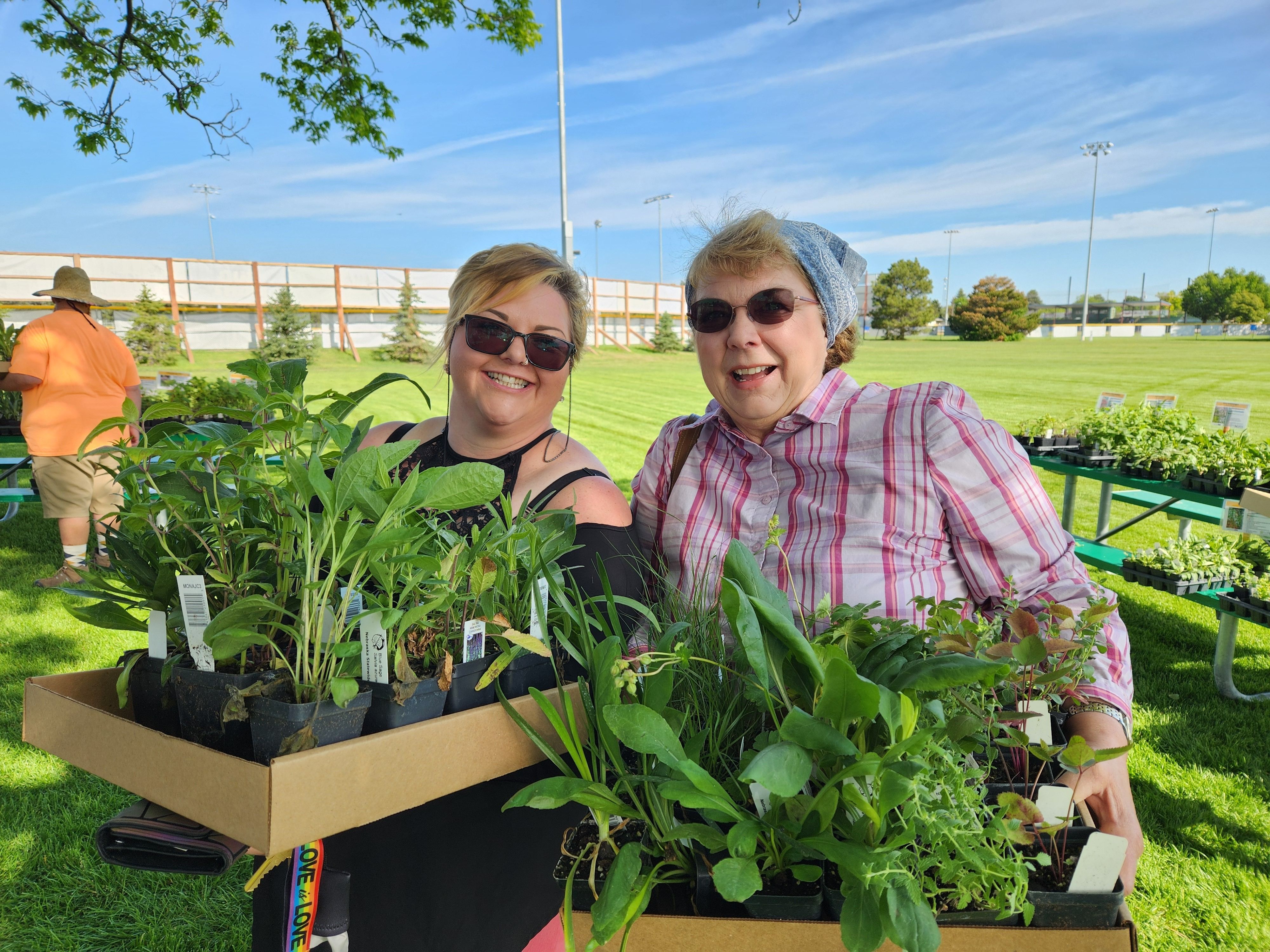 Two women hold flats of plants at an outdoor plant sale. 