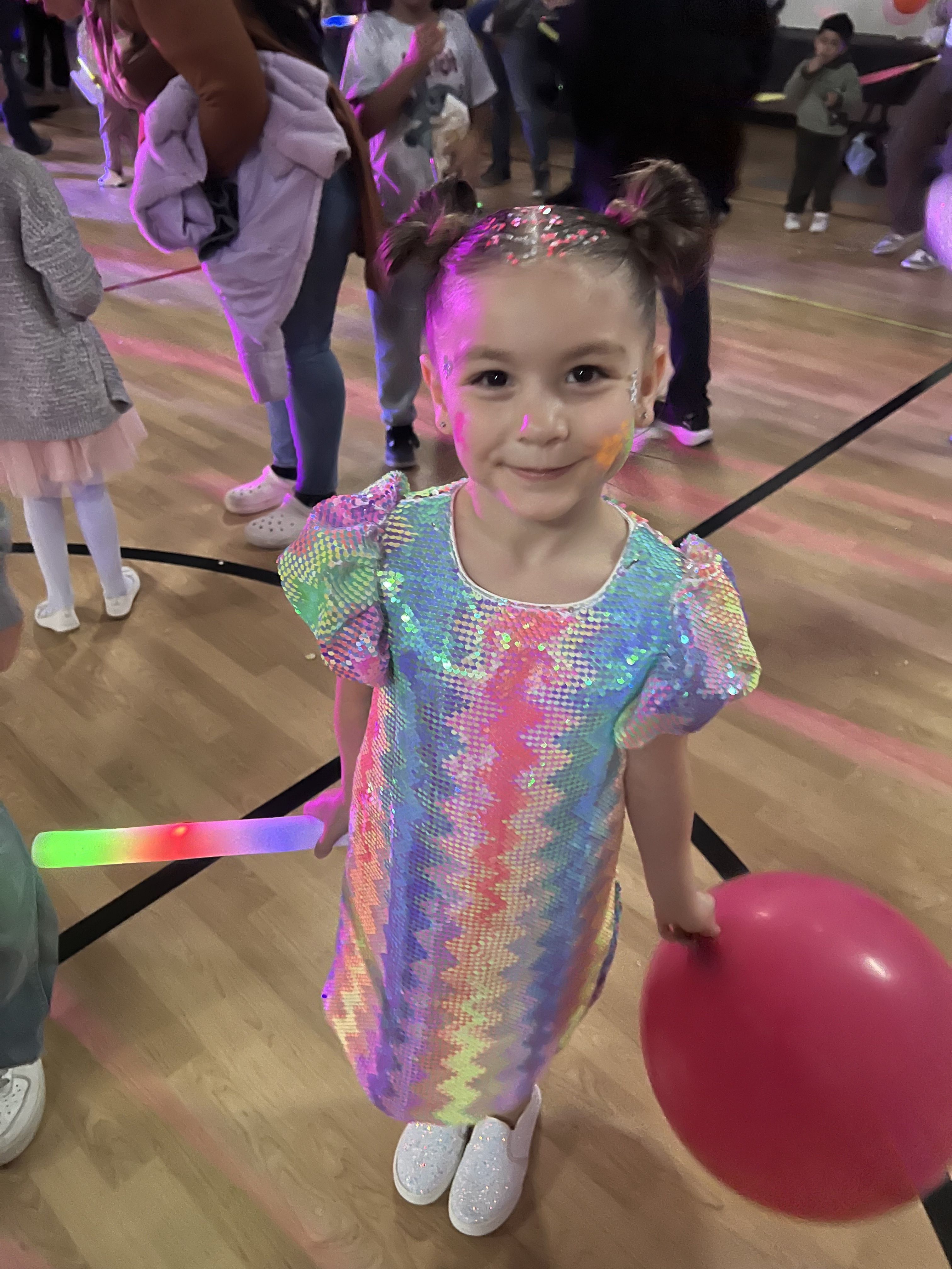 A young girl in a rainbow sequin dress smiles while holding a balloon at a school event, capturing joy, belonging, and the sparkle of possibility.