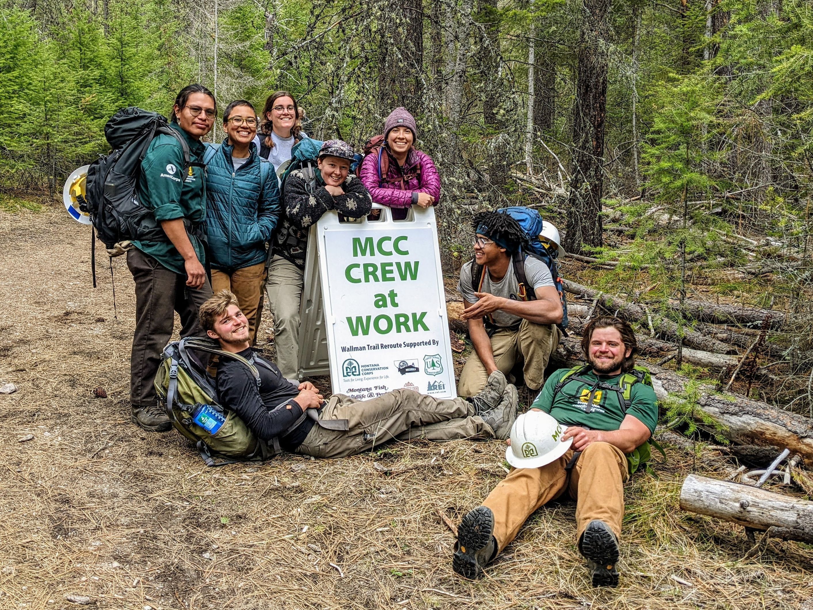 A crew stands smiling next to a sign that reads "MCC Crew At Work" and "Wallman Trail Sponsored By" with a list of logos and sponsors below.