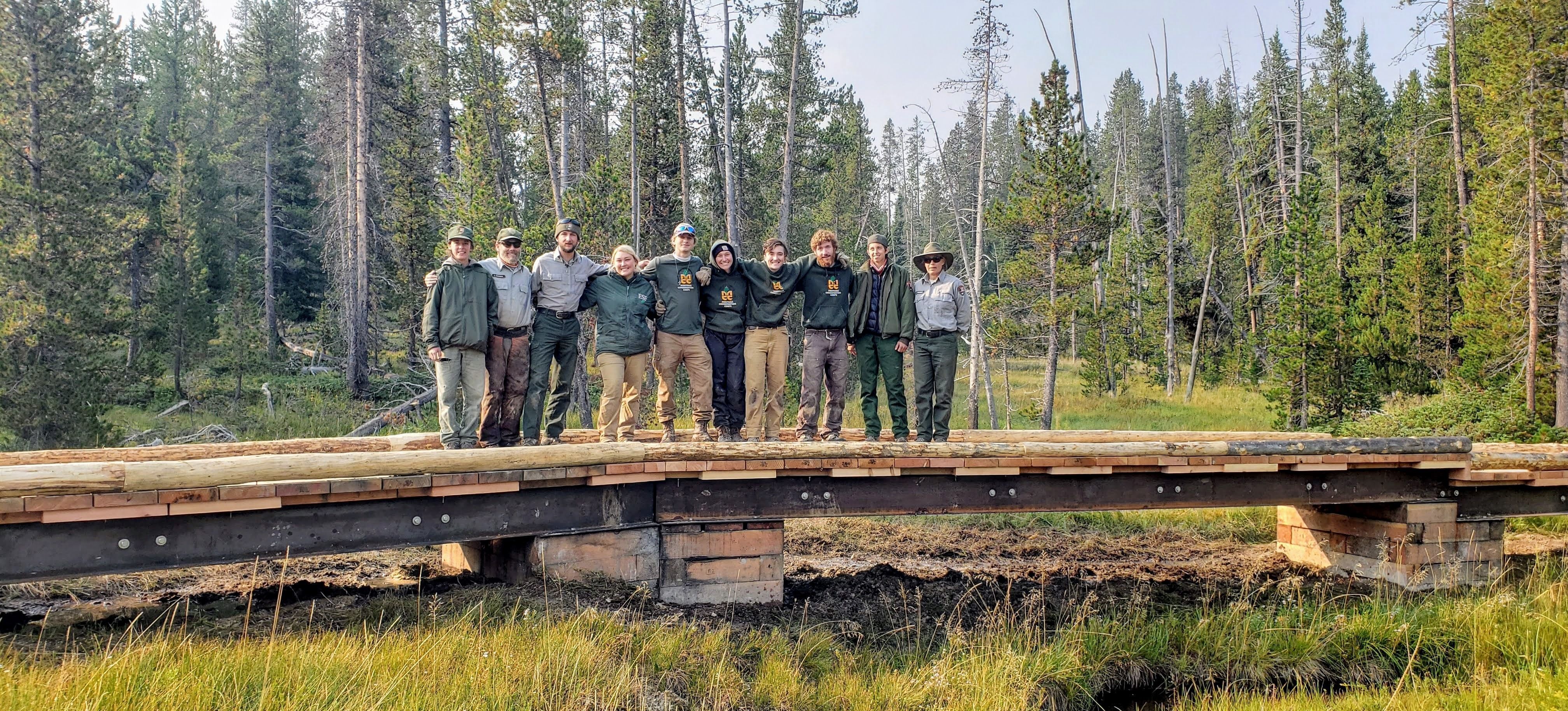 [Image Description: Eight MCC members and two Project Partners standing, arm in arm, on a newly built trail bridge, surrounded by tall trees.]