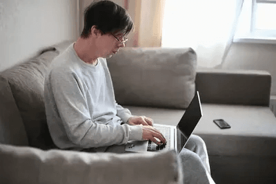 The image shows a person wearing glasses and casual gray clothing sitting on a gray couch while working on a laptop placed on their lap. The background includes a bright window and a smartphone resting on the couch beside them.