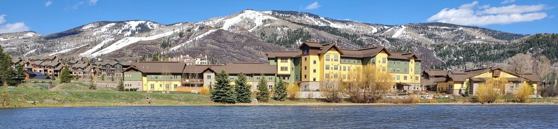 A lakeside building with mountains in the background, reflected in a calm lake under a blue sky.