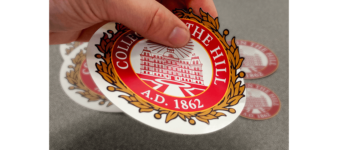 Hand showing a custom die cut decal with gold filigree edges - College on the Hill seal