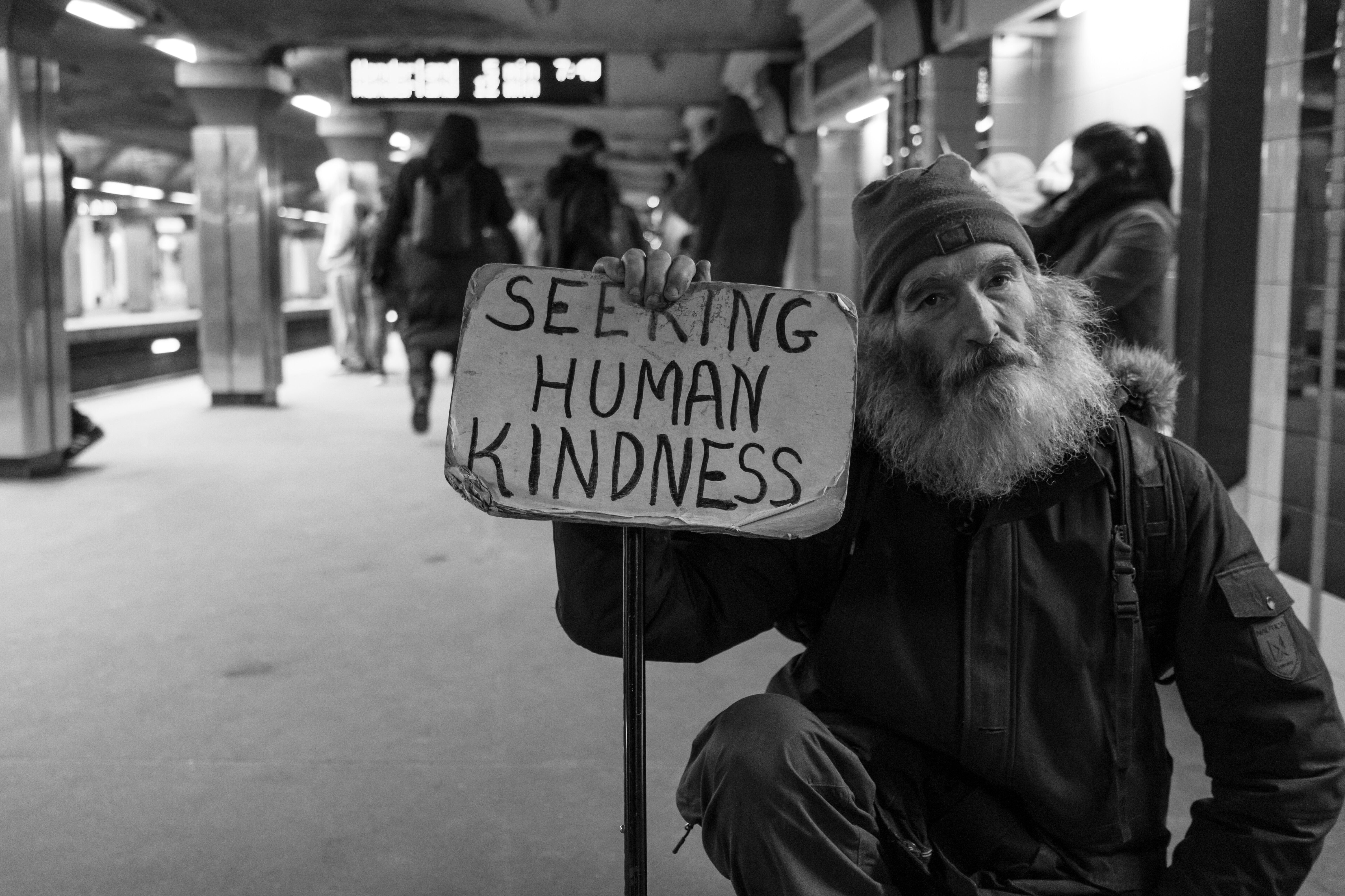 Picture of an older unhoused man, with a long beard holding a cardboard sign that says "seeking human kindness"