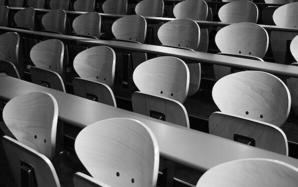 Photo of a series of auditorium desk chairs and long tables. The photo shows parts of three rows at an angle, so the chairs face slightly to the left.