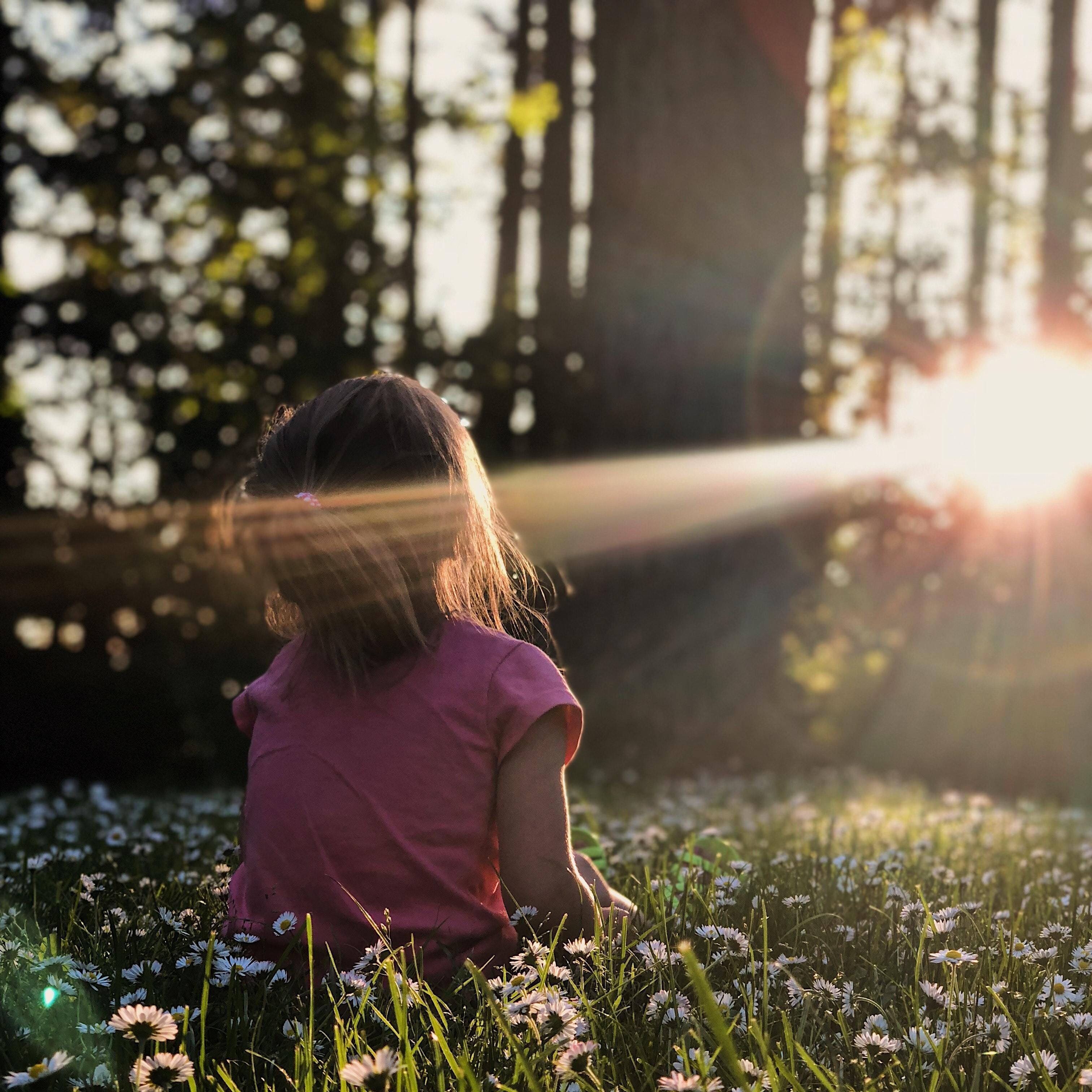 Image of a girl in a forest. She is sitting in a field of flowers, staring at the sun in between the trees.