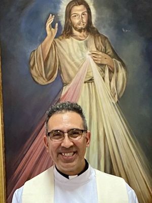 Smiling Hispanic priest in front of an image of the Divine Mercy