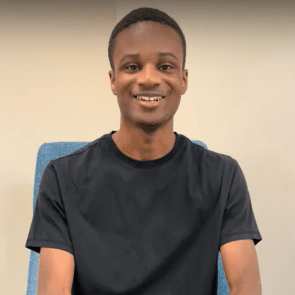 Jonathan is a Black man with short black hair. He wears a black t-shirt and sits on a blue chair, which peeks out over his shoulders. He smiles for the camera.