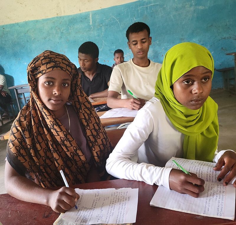Two 8th grade girls write in their notebooks during a Saturday Tutorial class, with classmates studying at desks behind them.